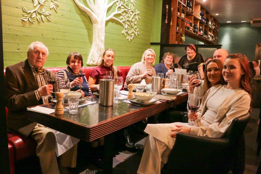 A full table of guests smiling in front of the olive tree wall